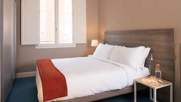 A neatly made bed with white linens and a red throw blanket, a bedside table with a lamp and a bottle, and a window with blinds.
