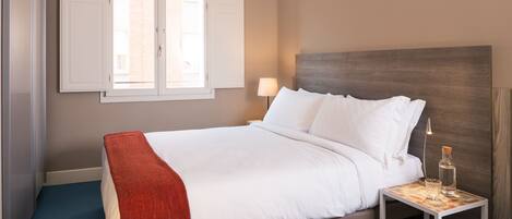 A neatly made bed with white linens and a red throw blanket, a bedside table with a lamp and a bottle, and a window with blinds.