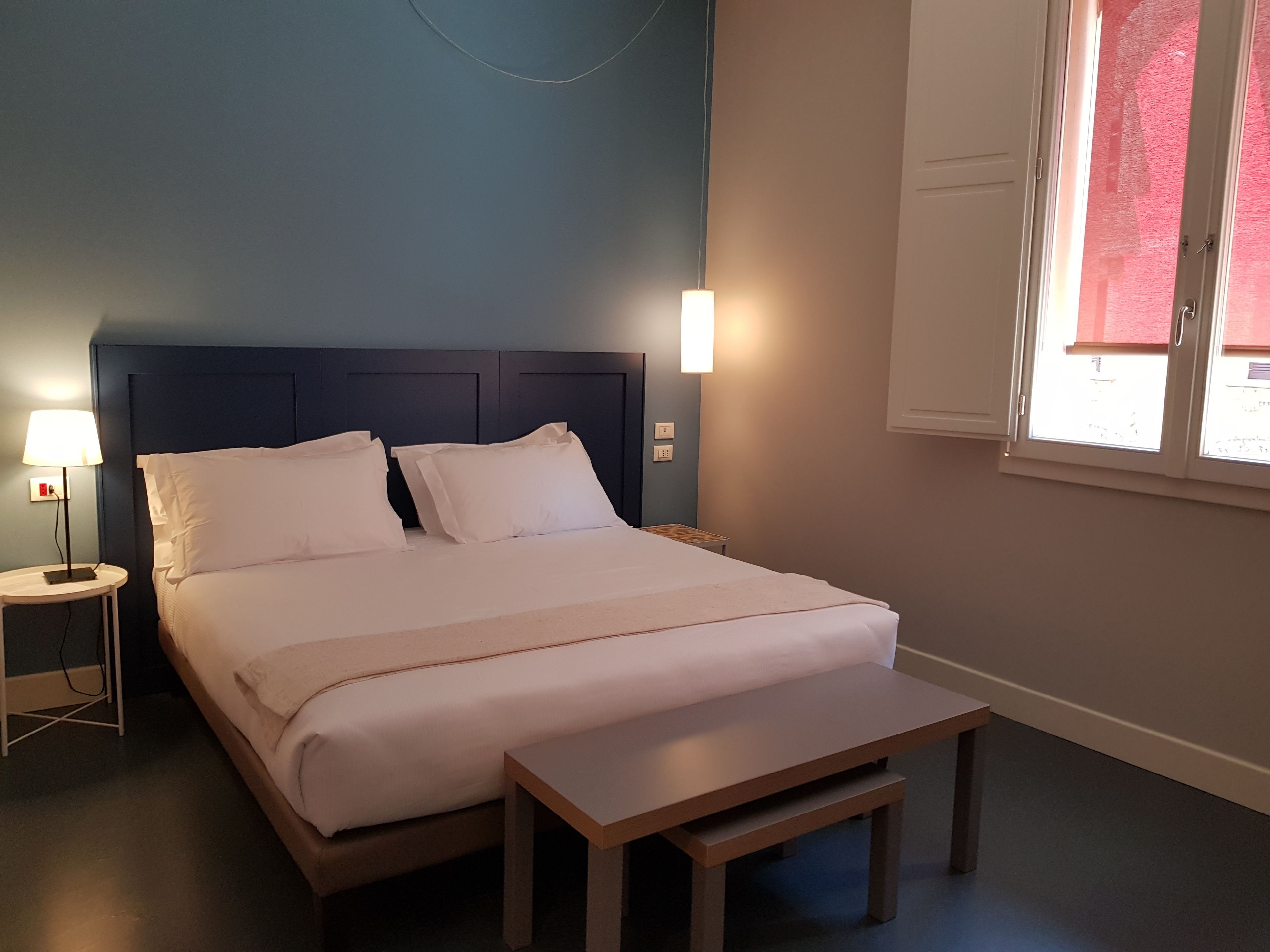 A neatly made bed with white linens, a bedside table with a lamp, and a window with blinds.