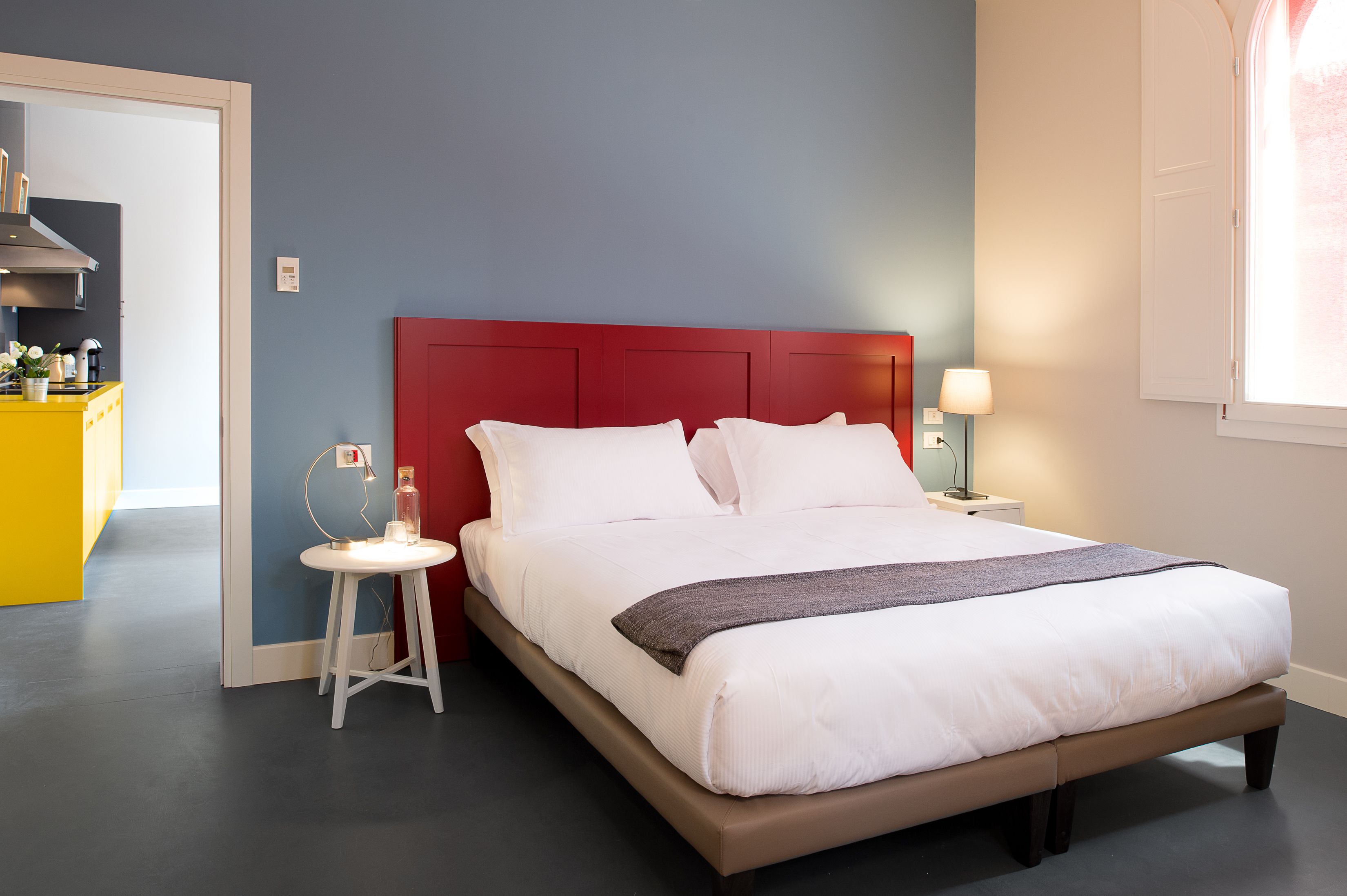 Un letto rifatto con lenzuola bianche e una testiera rossa, un comodino con una lampada e un piccolo sgabello.