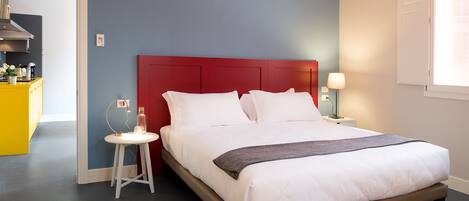 A neatly made bed with white linens and a red headboard, a bedside table with a lamp, and a small stool.