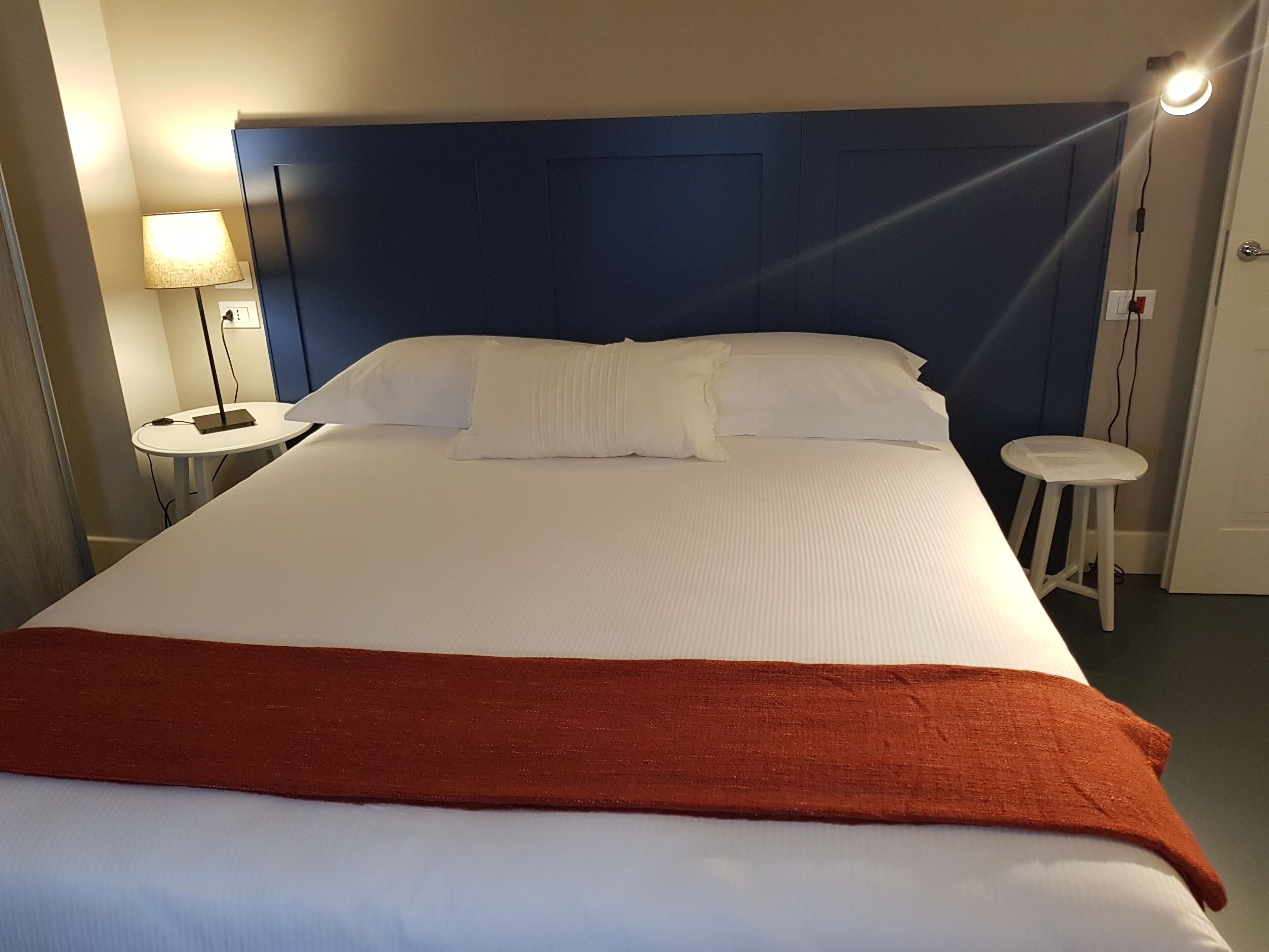 A hotel room with a large bed, a nightstand, a lamp, and a small stool.