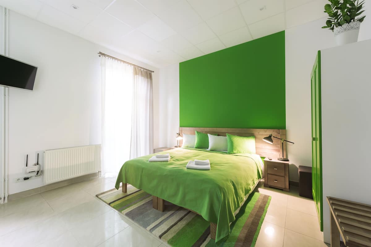 A modern hotel room with a green accent wall, a large bed with green bedding, a flat-screen TV mounted on the wall, a radiator, and a green-striped rug.