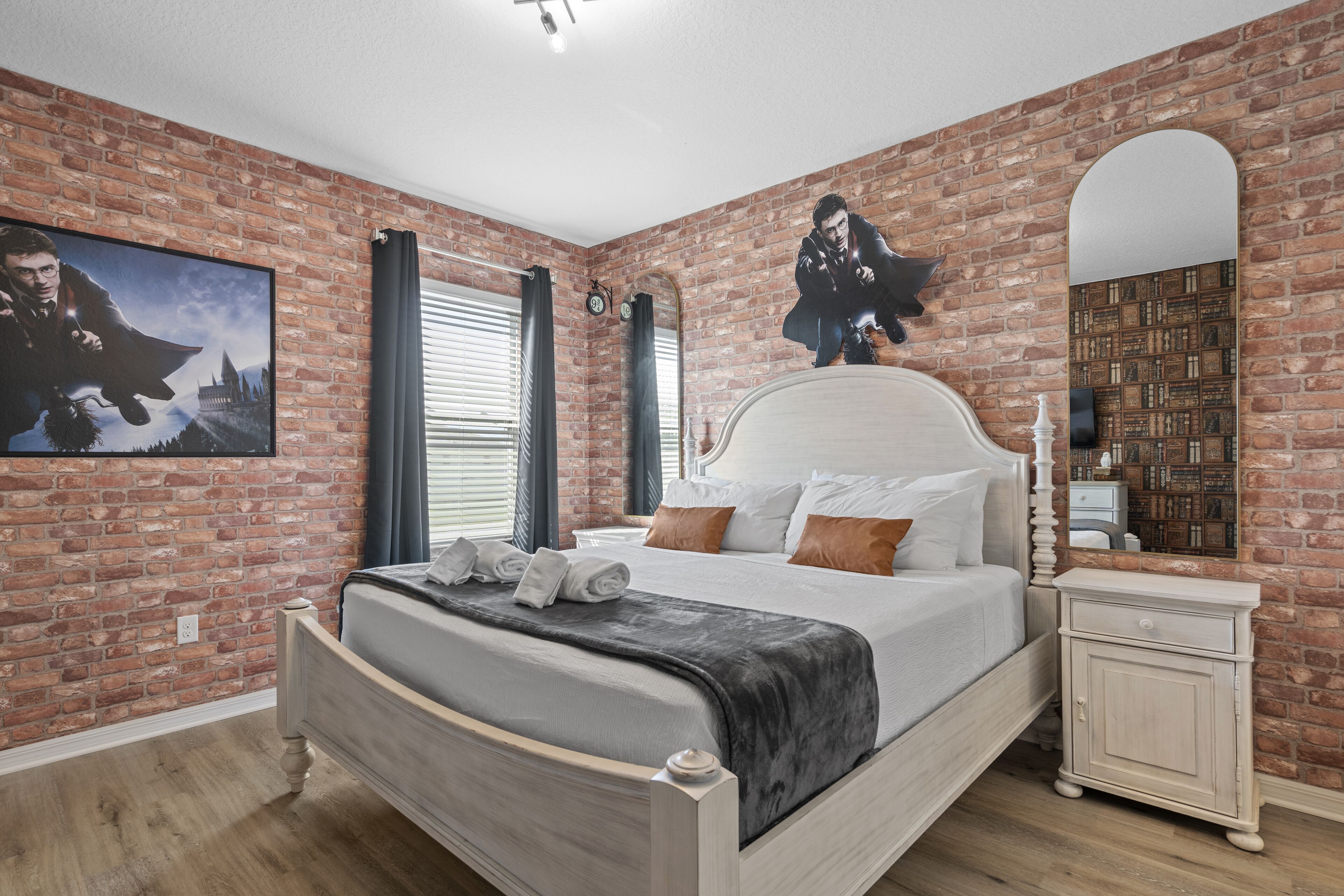 A bedroom with a brick wall, a large bed, a nightstand, a mirror, and a bookshelf.