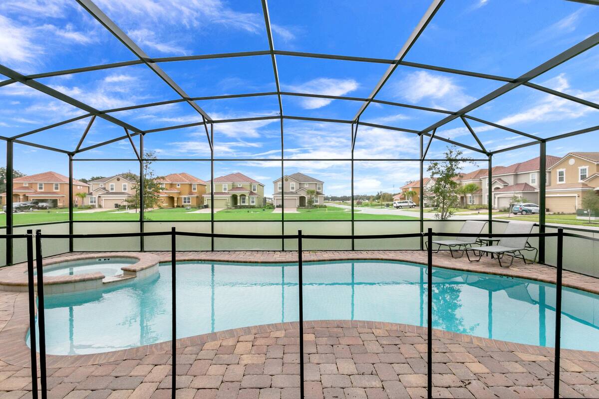 A covered outdoor swimming pool with a brick patio, surrounded by a metal fence, and houses in the background.