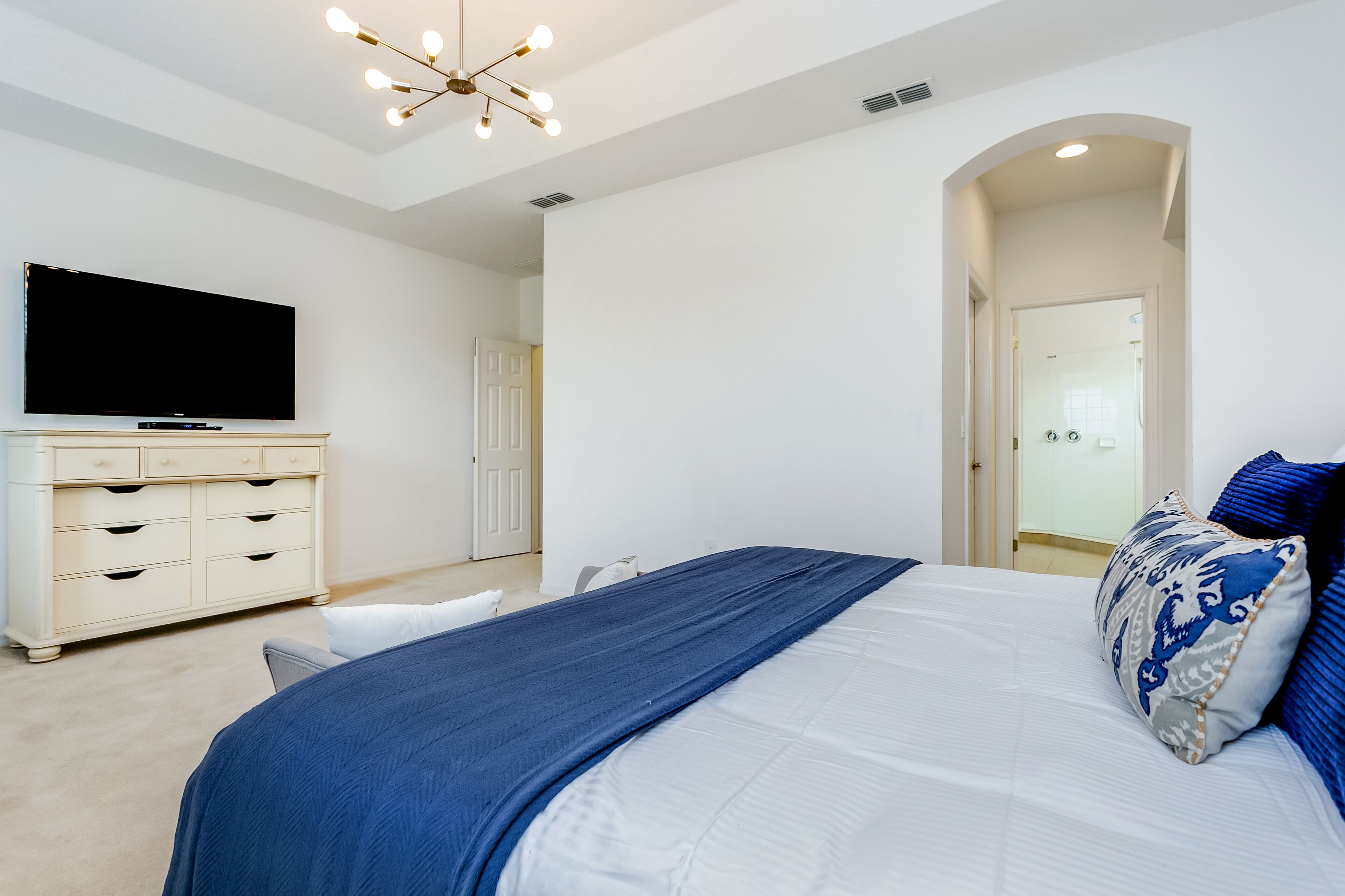 A bedroom with a large bed, a TV, a dresser, and a door leading to another room.