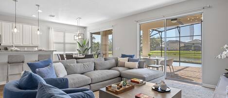 A spacious living room with a large sectional sofa, a coffee table with decorative items, and a view of the outdoors through large sliding glass doors.