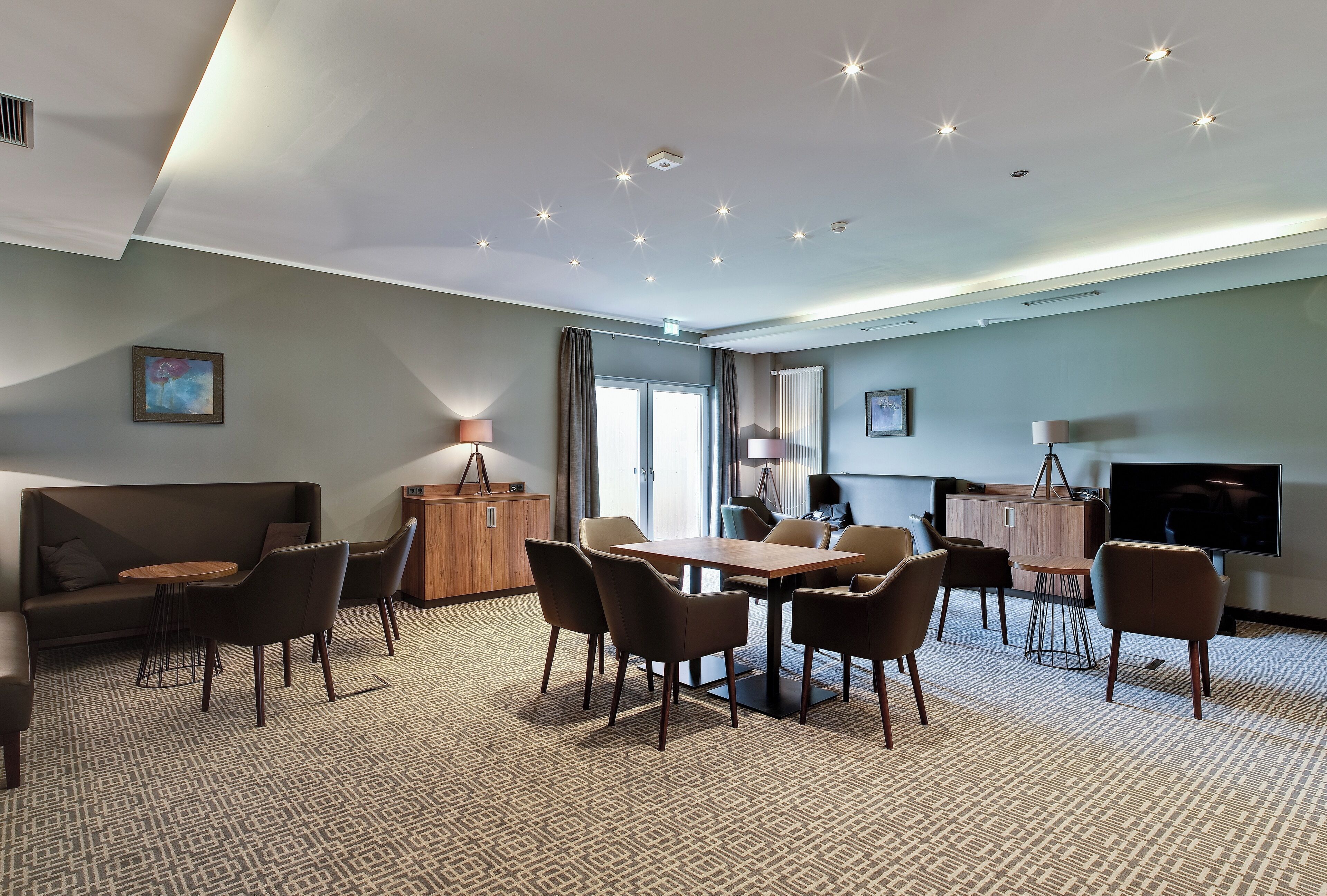 A modern hotel room with a dining area, a sofa, a TV, and a patterned carpet.