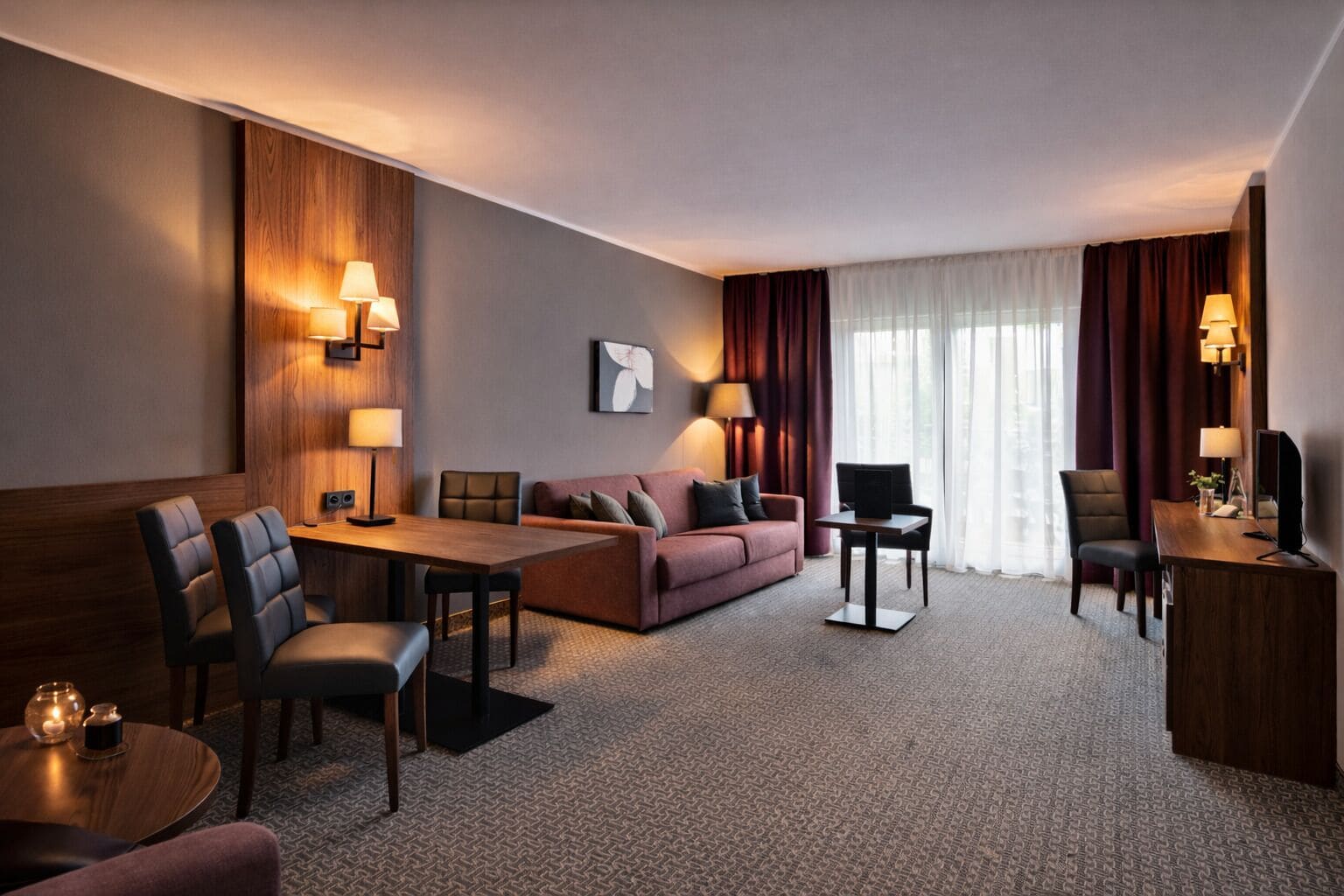Habitación de hotel con una pared de madera decorativa, un sofá, una mesa de comedor con sillas, un televisor y un escritorio con lámpara.