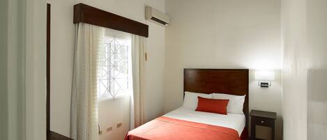 A bedroom with a bed, a nightstand, a window with curtains, and an air conditioning unit.