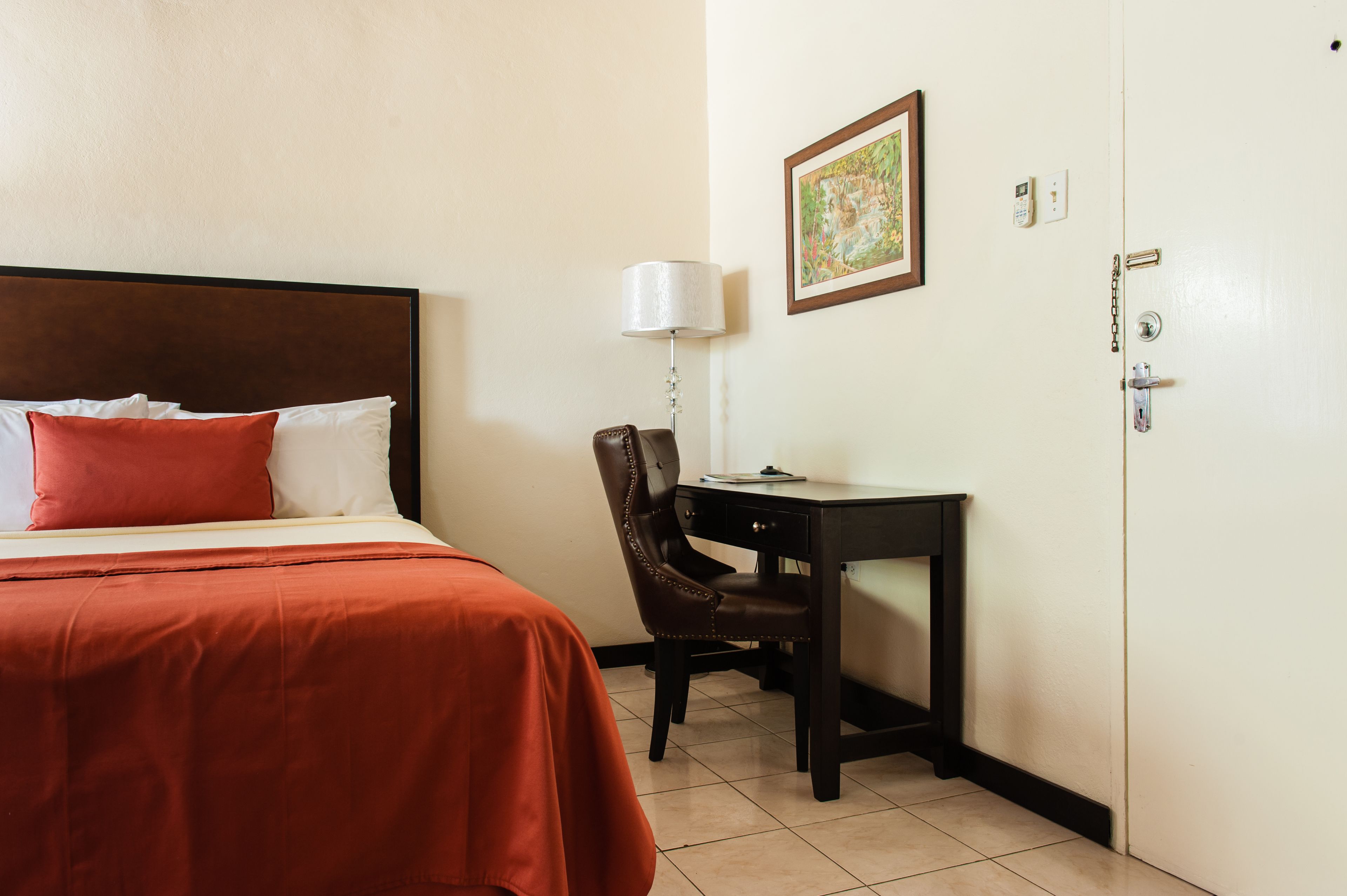 A hotel room with a bed, a desk, a chair, a lamp, a door, and a wall-mounted painting.