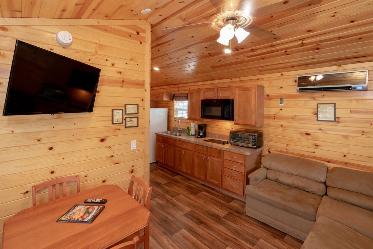 A wooden cabin interior with a dining table, chairs, a sofa, a kitchen area with a microwave, and a ceiling fan.