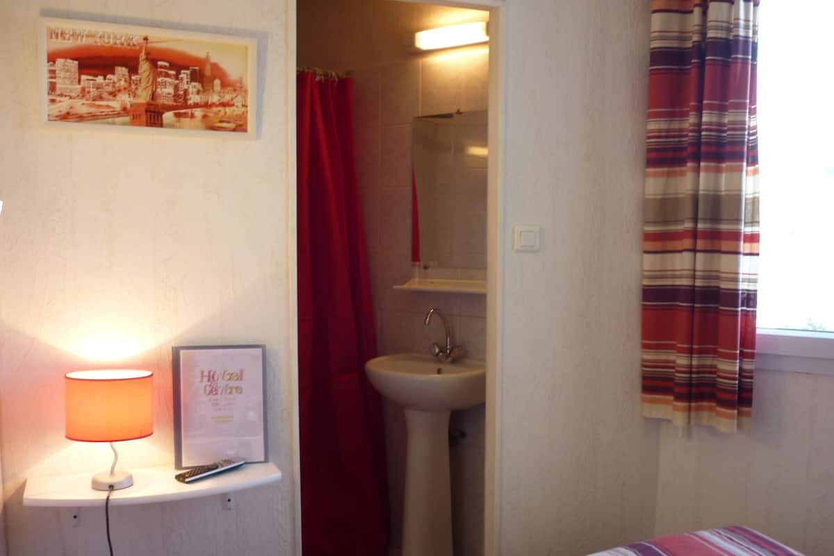 A hotel room with a bed, a nightstand with a lamp, a framed sign, a bathroom with a sink and mirror, and a window with curtains.