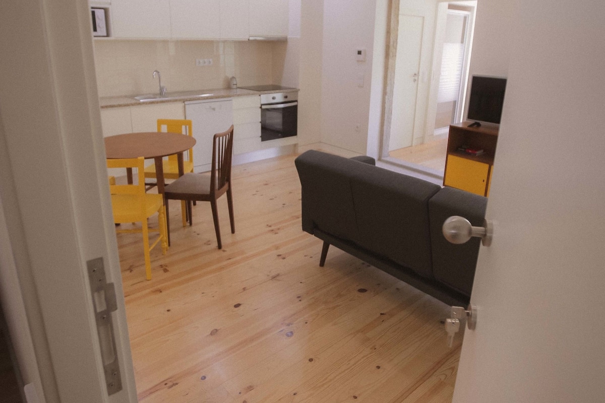 A compact kitchen with white cabinets, a dining table with chairs, and a gray sofa in a hallway.