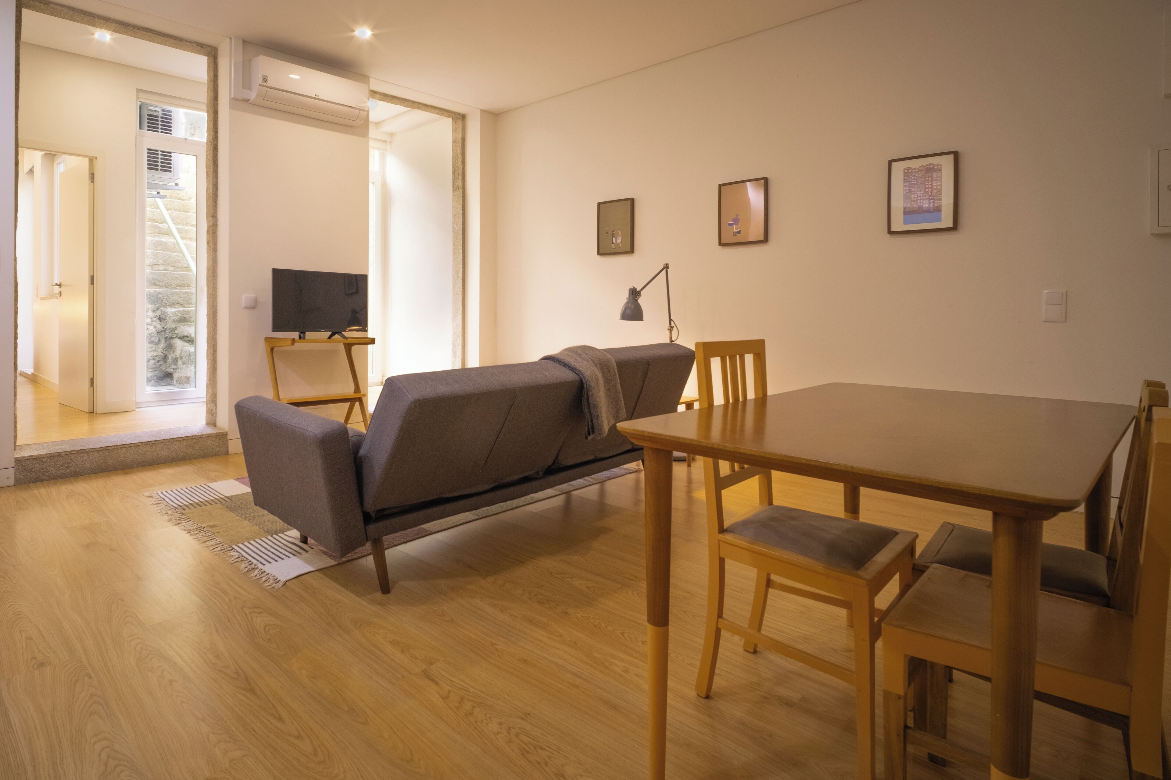 A modern living room with a sofa, a dining table, and chairs. There is a TV on a stand, a lamp, and framed pictures on the wall.