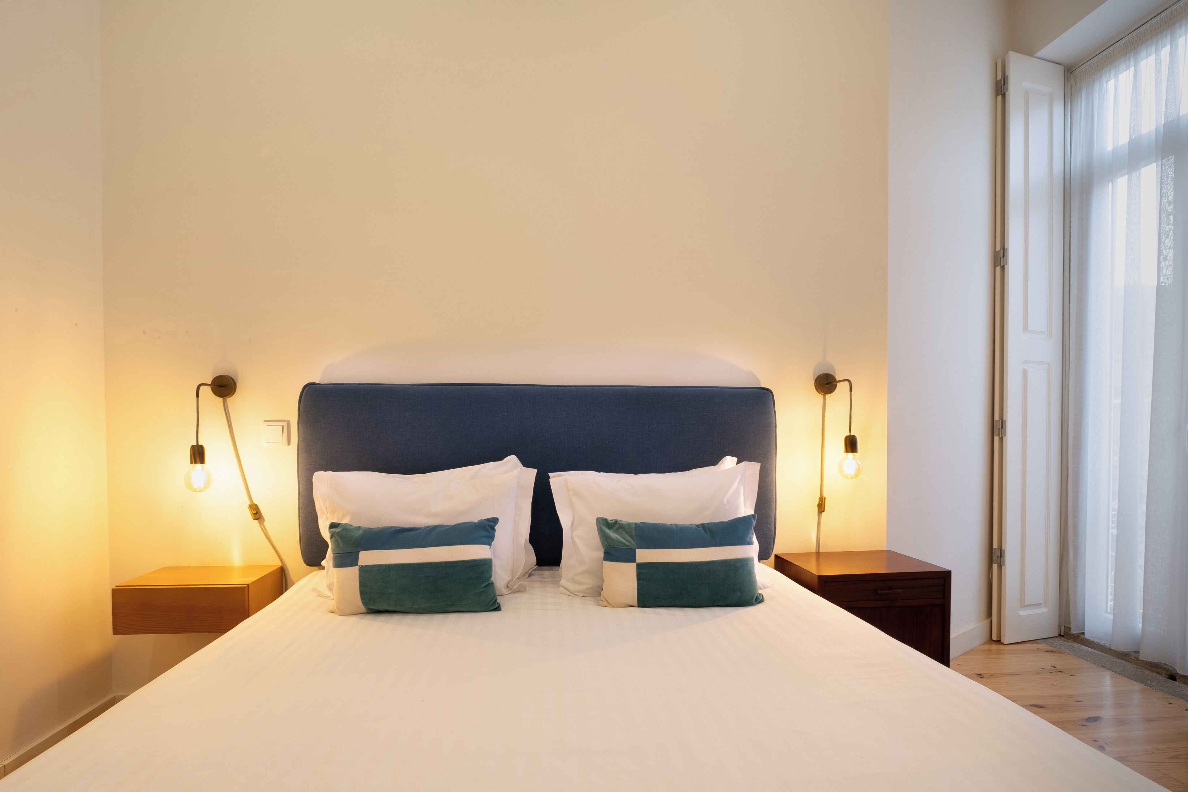 A neatly made bed with white and teal pillows, two bedside tables with lamps, and a large window with curtains.