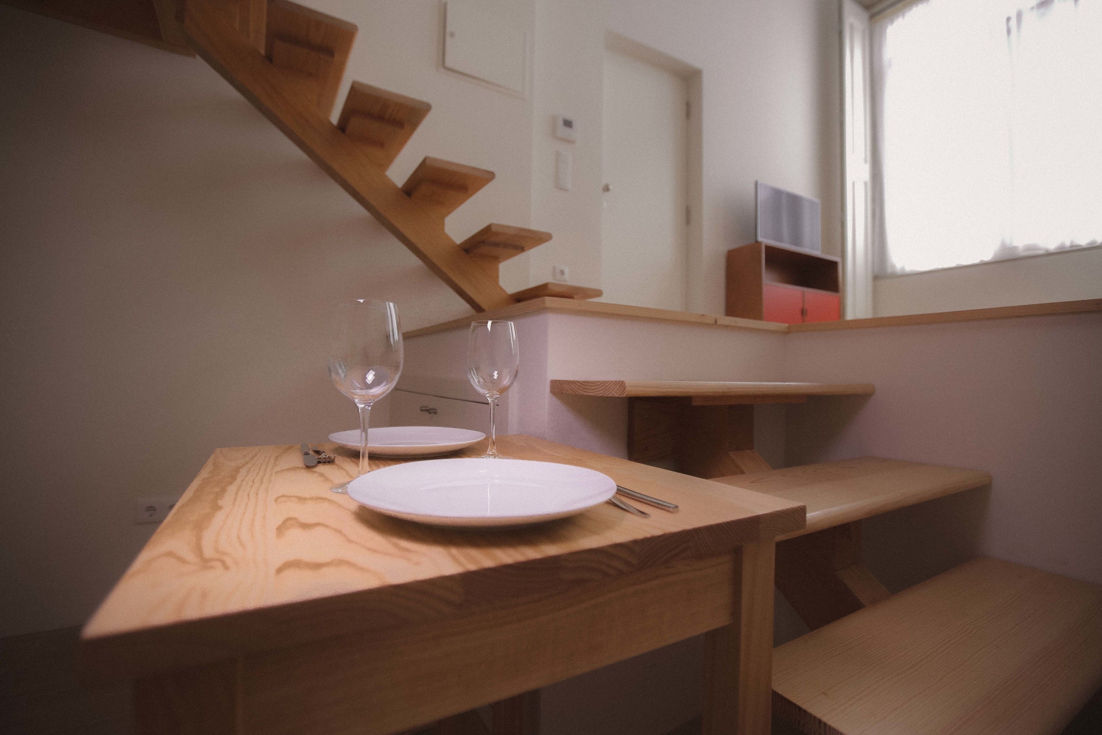 Une table de salle à manger en bois, avec deux assiettes blanches et deux verres à vin, un banc en bois et un escalier avec une rampe en bois.