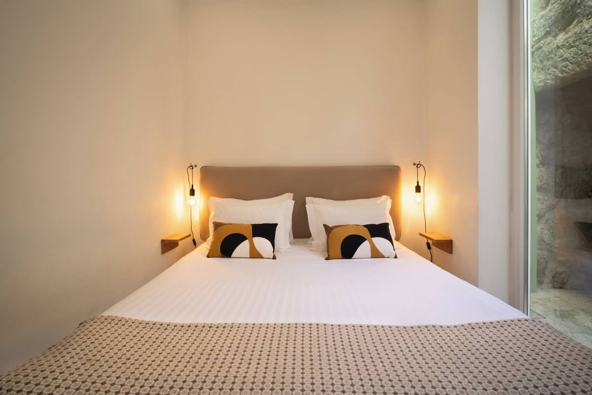 A neatly made bed with patterned bedding, two wall-mounted reading lamps, and a view of a stone wall through a window.