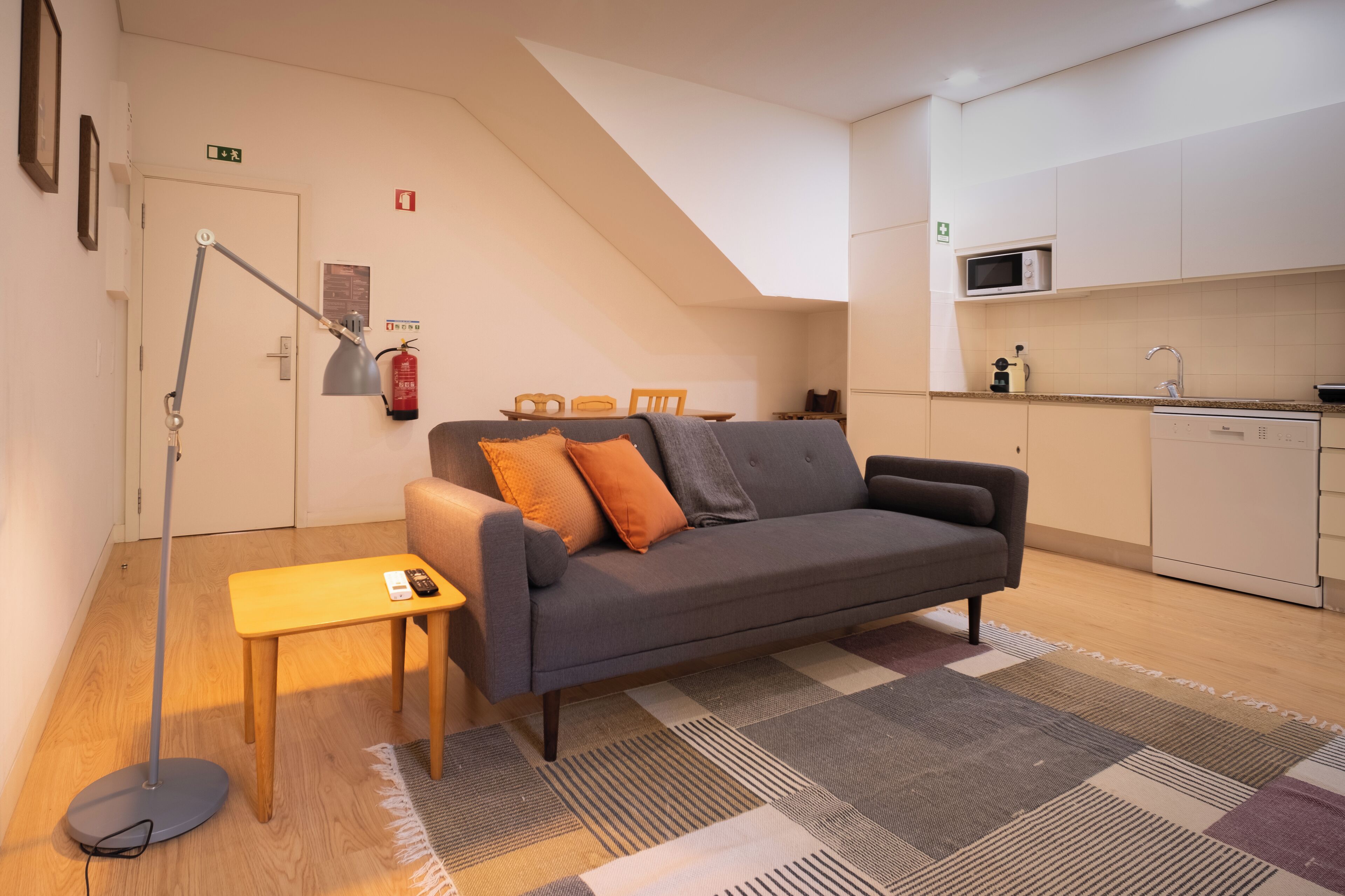 A modern living room with a sofa, a small table, a floor lamp, and a kitchen area with a microwave and sink.