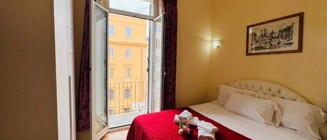 A hotel room with a bed, a window with curtains, a painting on the wall, and a view of buildings outside.