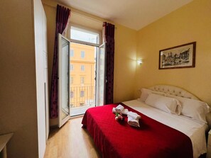 Hotellihuone, jossa on sänky, ikkuna verhoilla, taulu seinällä ja näkymä ulkona oleviin rakennuksiin.