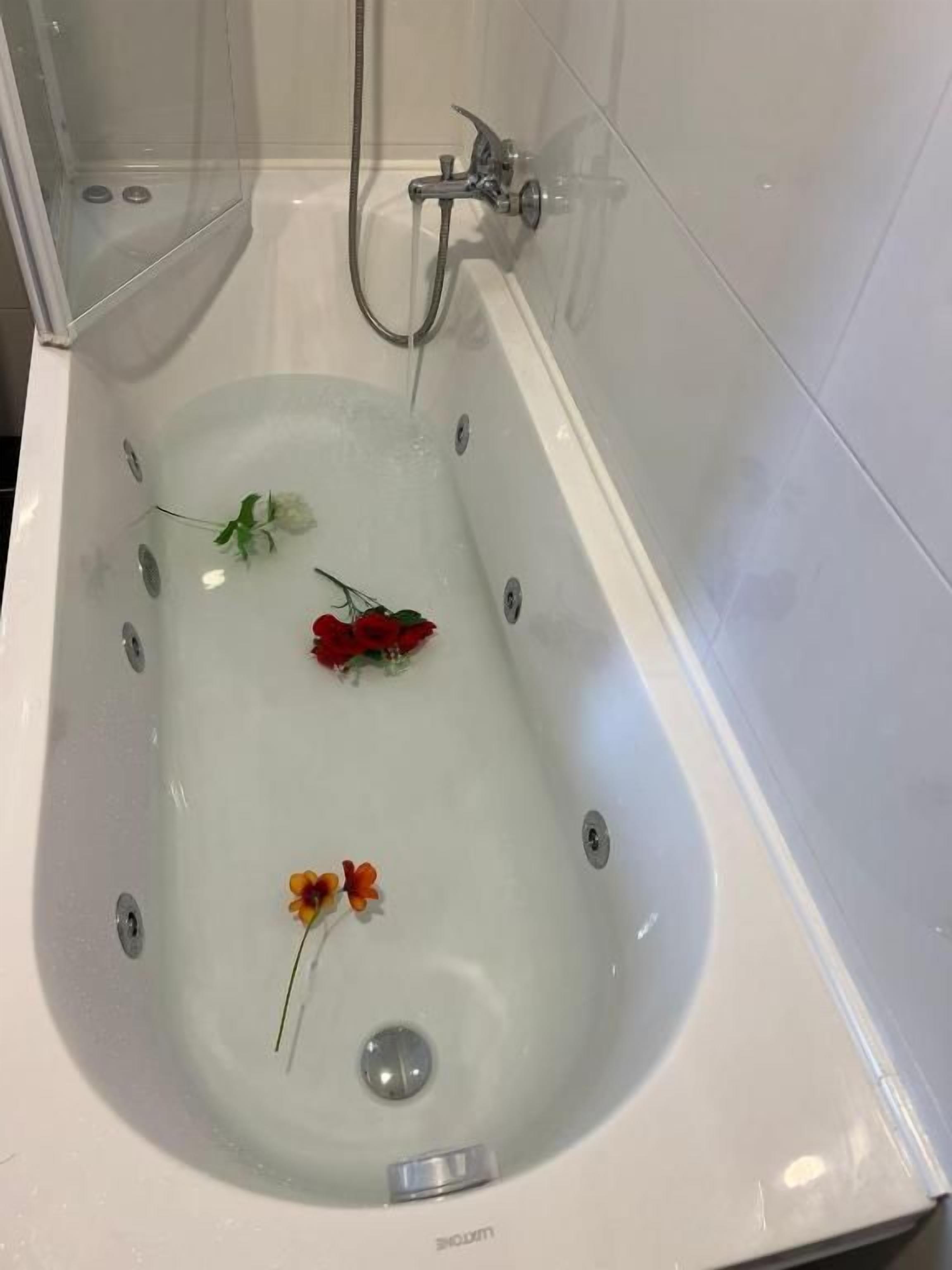 A bathtub with a shower head, a clear glass enclosure, and a single flower floating on the water.
