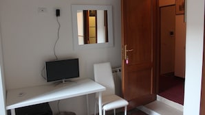 Una habitación con un escritorio blanco, una computadora, una silla, una puerta de madera y un espejo en la pared.