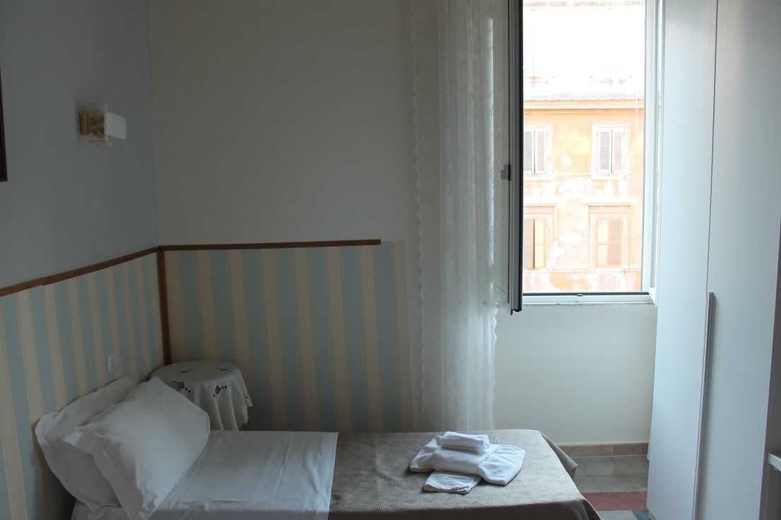 Une petite chambre avec un lit simple, une tête de lit à rayures, une fenêtre avec des rideaux et une vue sur les bâtiments environnants.
