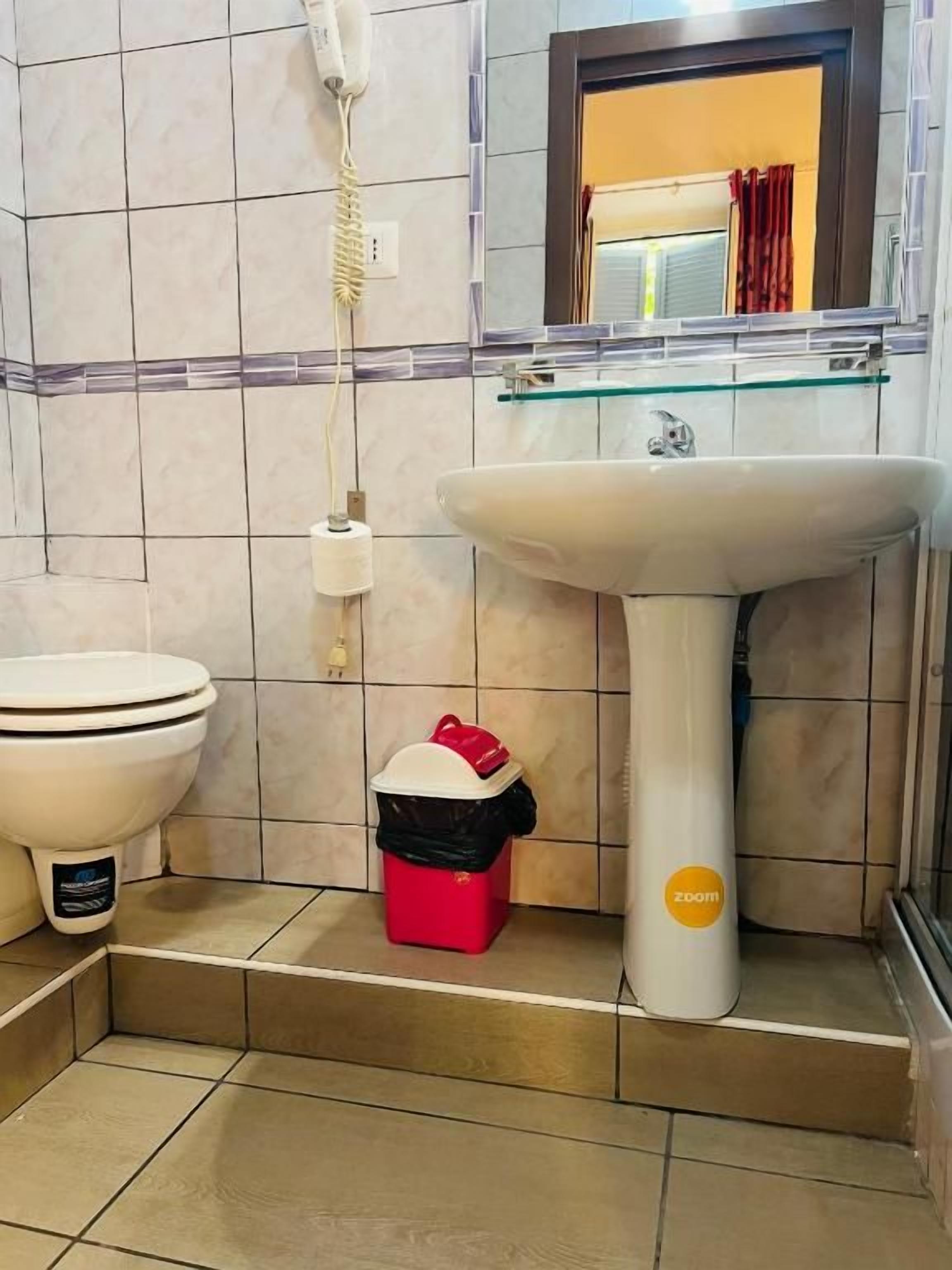 Un baño con un lavamanos pedestal blanco, un inodoro, un cubo de basura rojo y un teléfono fijo en la pared.