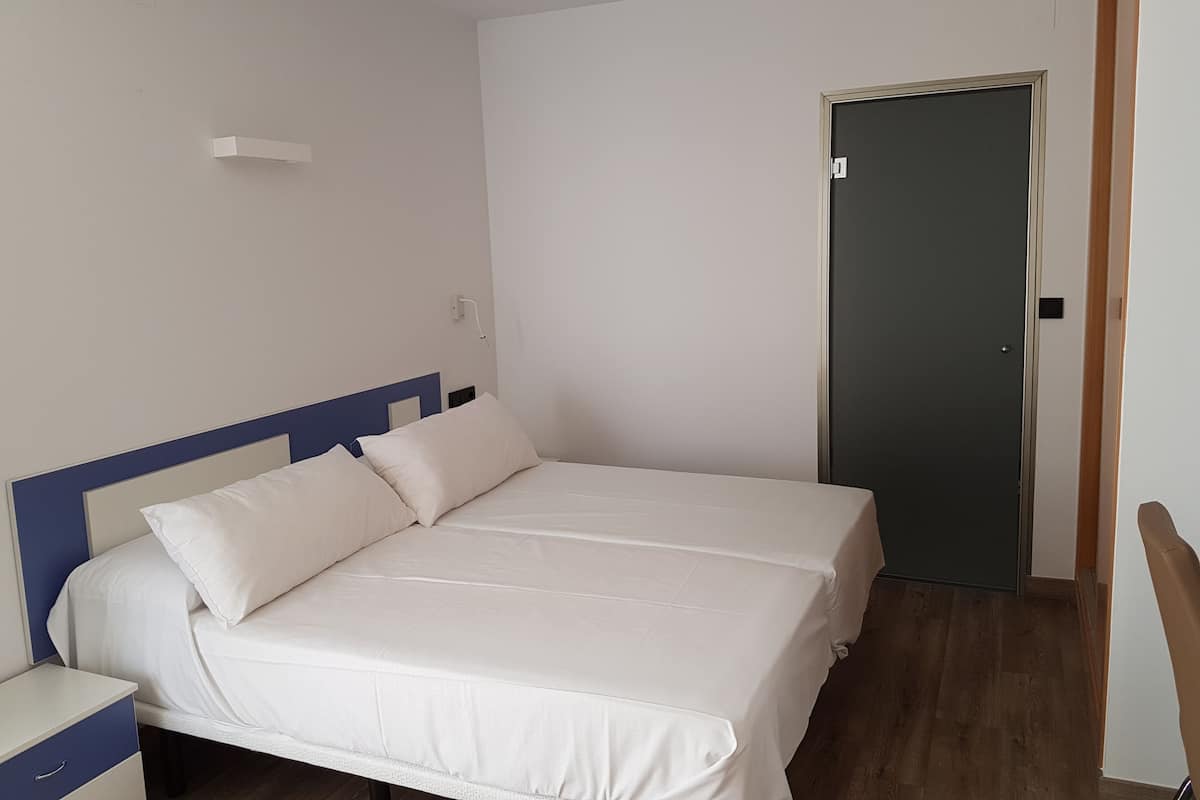 Un lit bien fait, avec du linge de lit blanc et une tête de lit bleue, dans une pièce aux murs blancs et au plancher en bois.