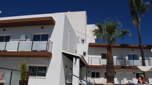 Ein modernes Hotel mit heller Fassade, mehreren Balkonen und einem Poolbereich mit Liegestühlen und Palmen.