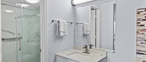 Un baño moderno con cabina de ducha de vidrio, un lavamanos blanco con espejo encima y un perchero con toallas blancas.