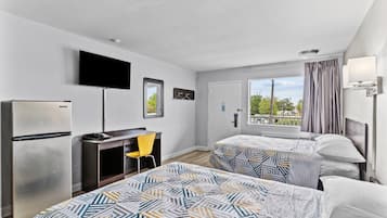 Habitación de hotel con dos camas, un escritorio, un refrigerador, un televisor montado en la pared y una ventana con cortinas.