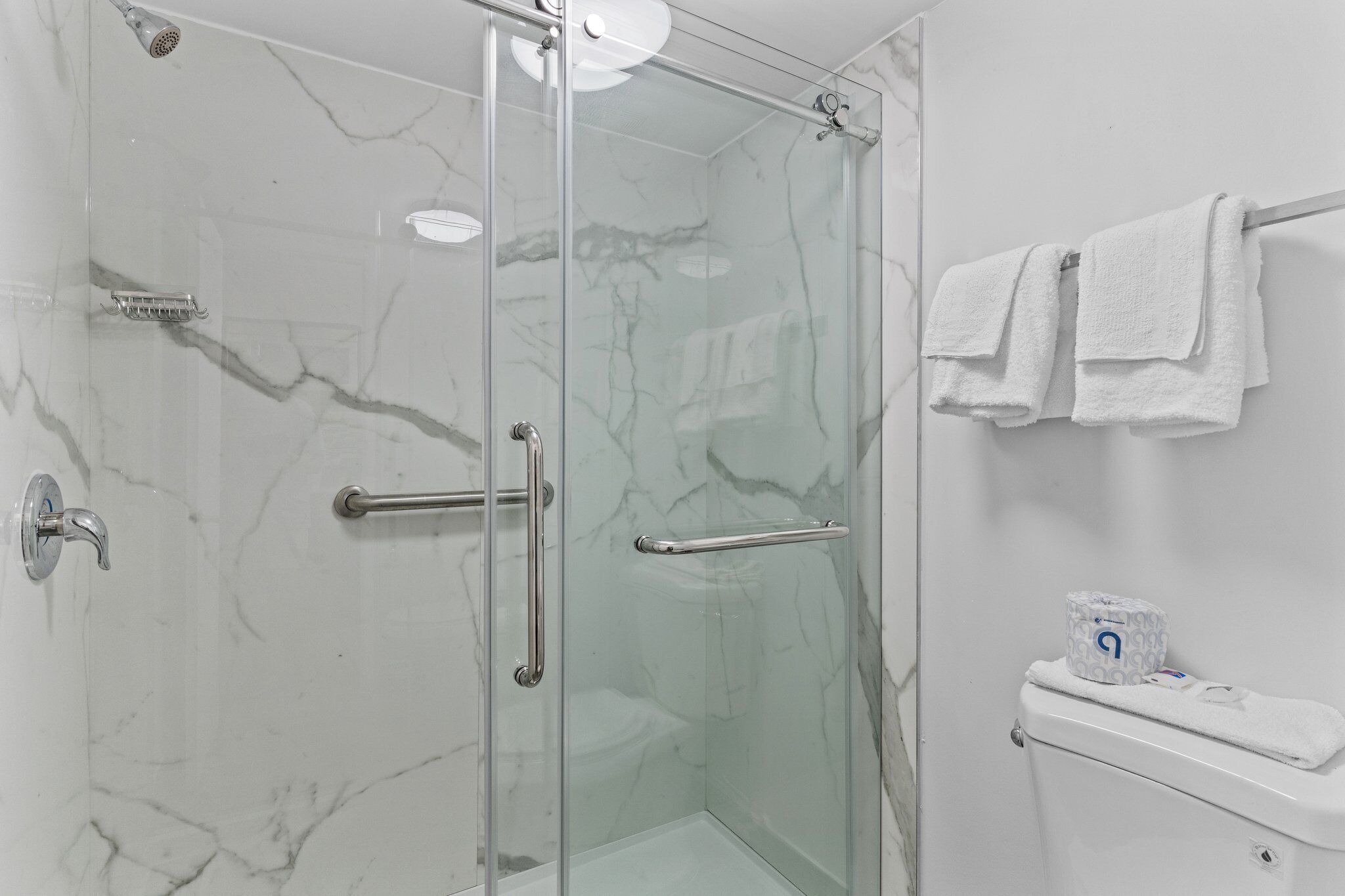 A modern bathroom with a glass-enclosed shower, marble walls, a toilet, and two towels hanging on a rack.