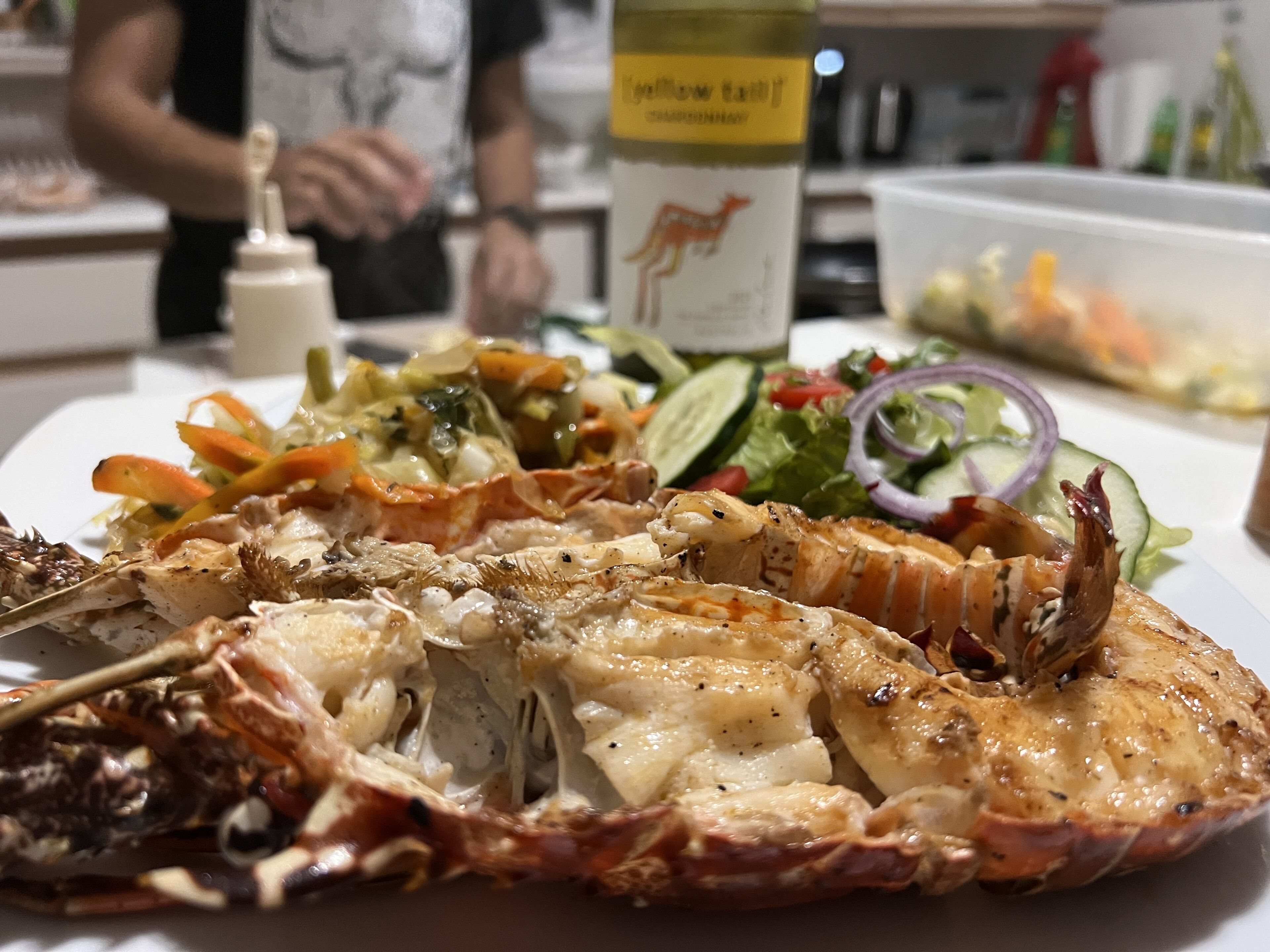Une queue de homard présentée sur une assiette, accompagnée d’une salade, et une bouteille de vin en arri ère-plan.