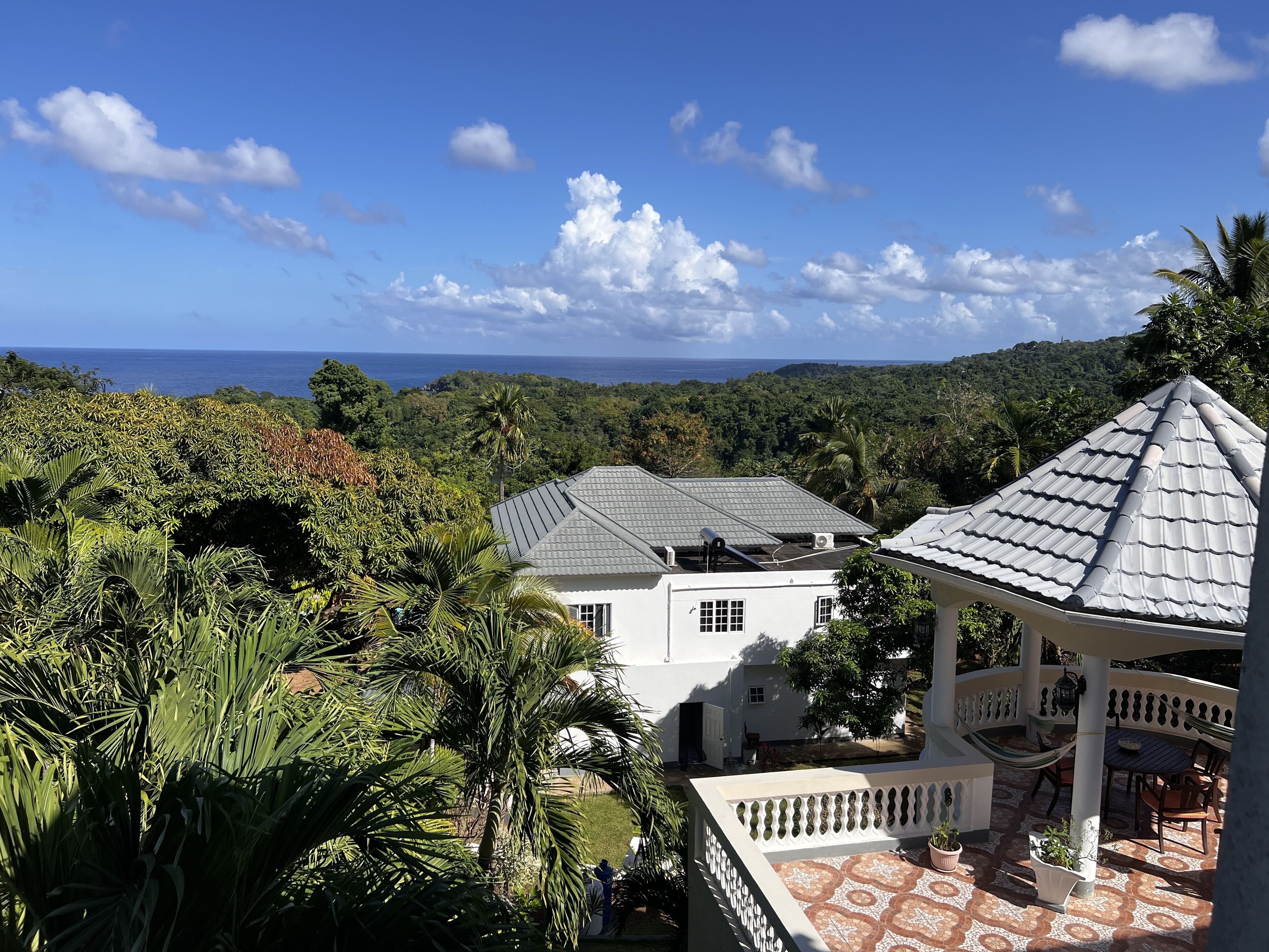 A white building with a grey roof, surrounded by lush greenery and a view of the ocean.