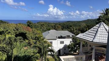A white building with a grey roof, surrounded by lush greenery and a view of the ocean.