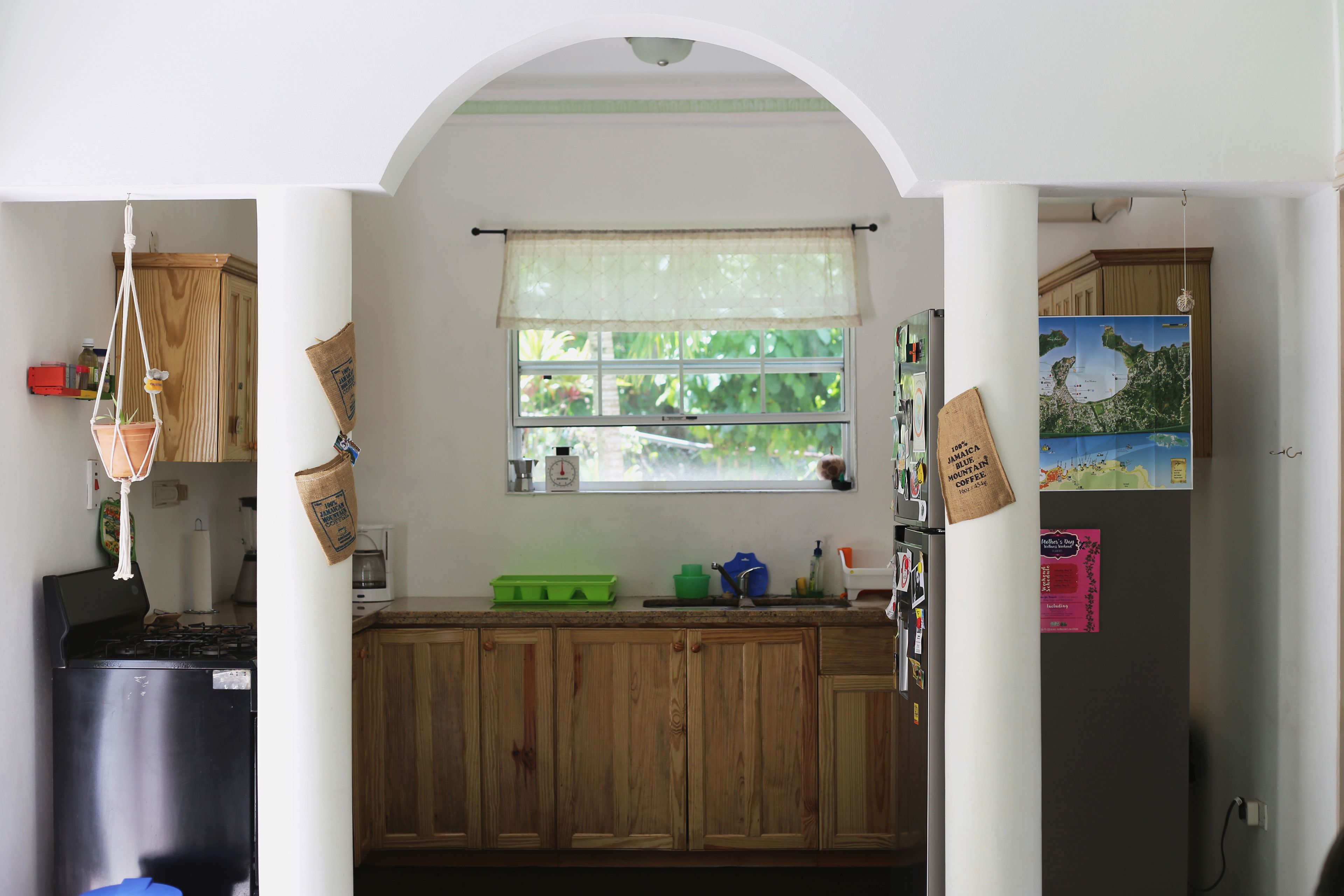 A kitchen with wooden cabinets, a window with a sheer curtain, a green tray, and a refrigerator with magnets and notes.