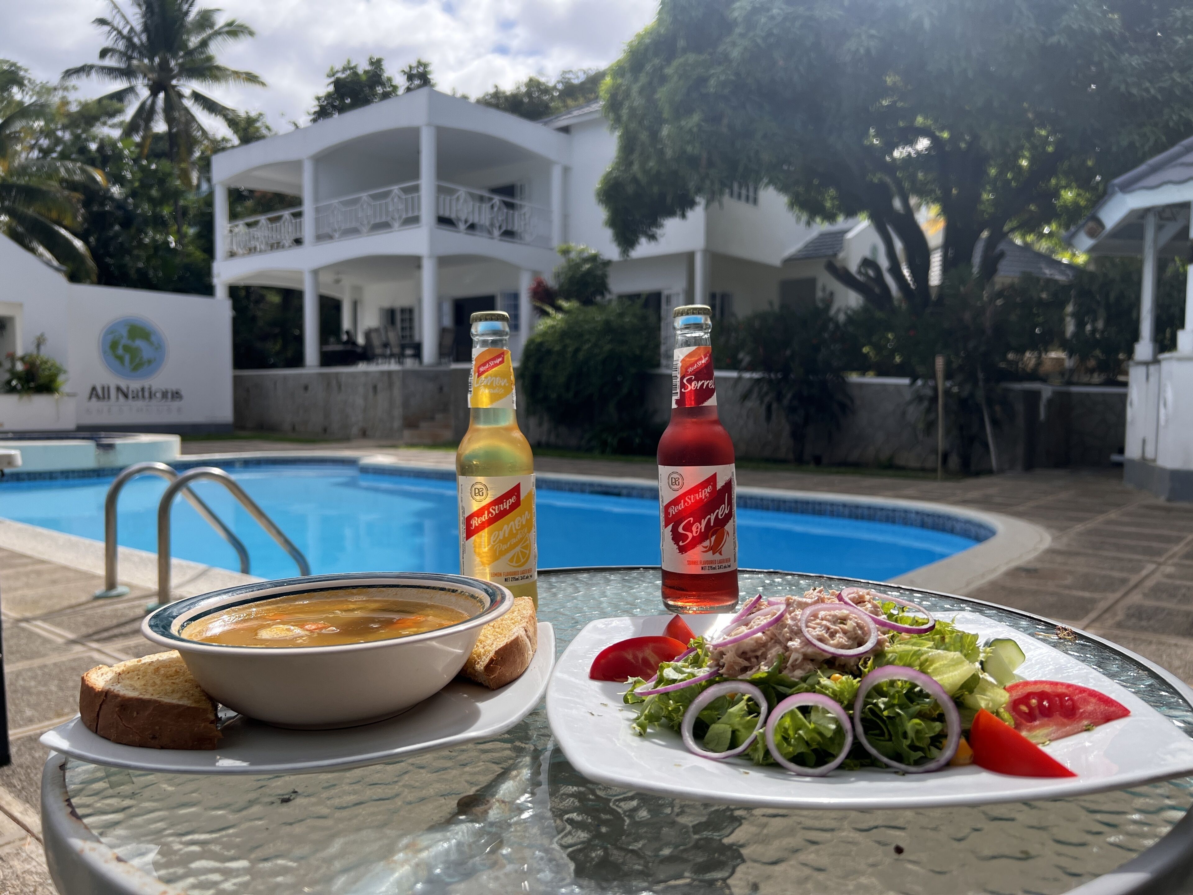 A dining table with a bowl of soup, a plate of salad, and two bottles of Red Stripe beer by a poolside.