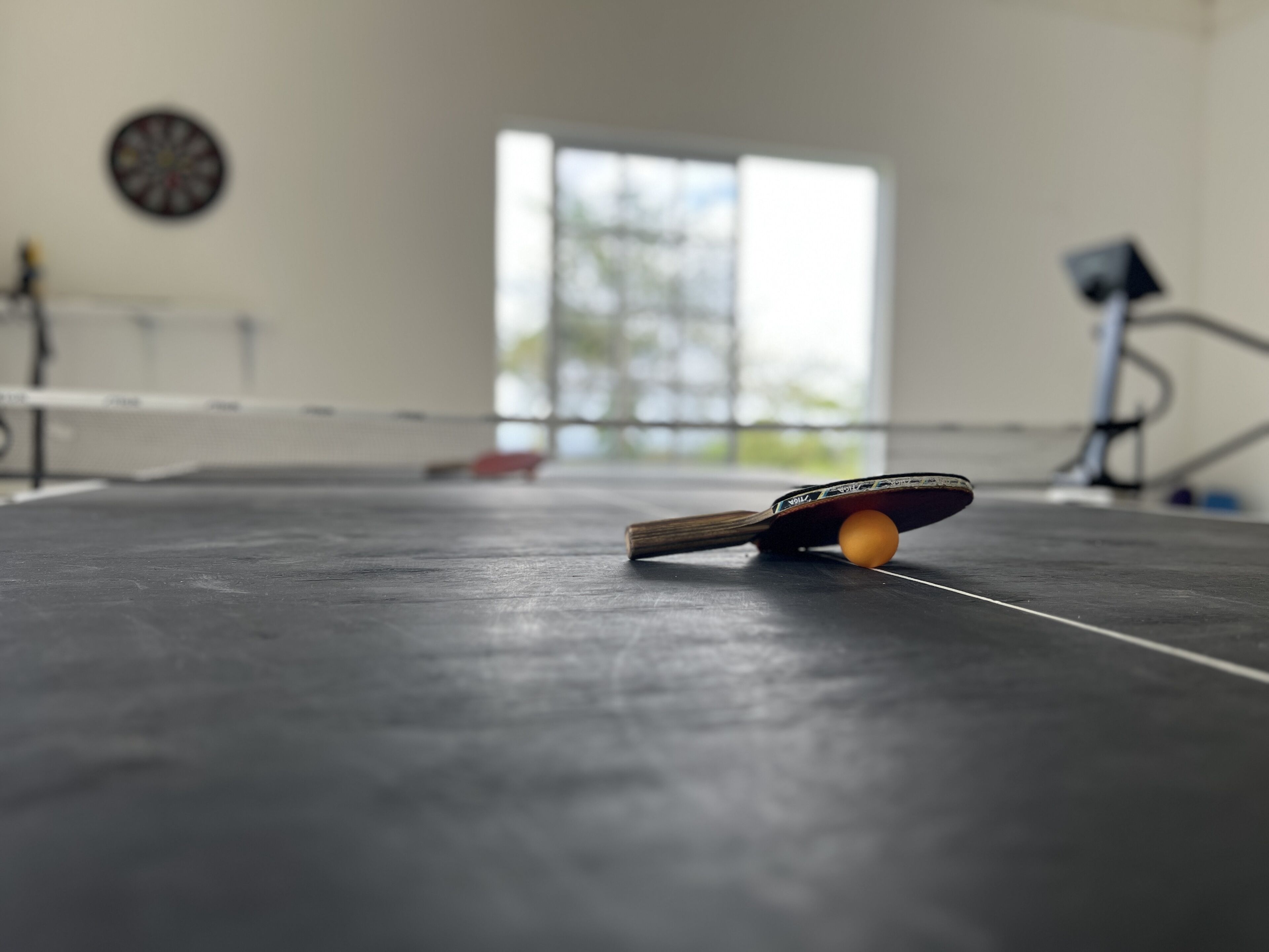 A table tennis table with a ball and paddle in the foreground, a dartboard on the wall, and a window in the background.