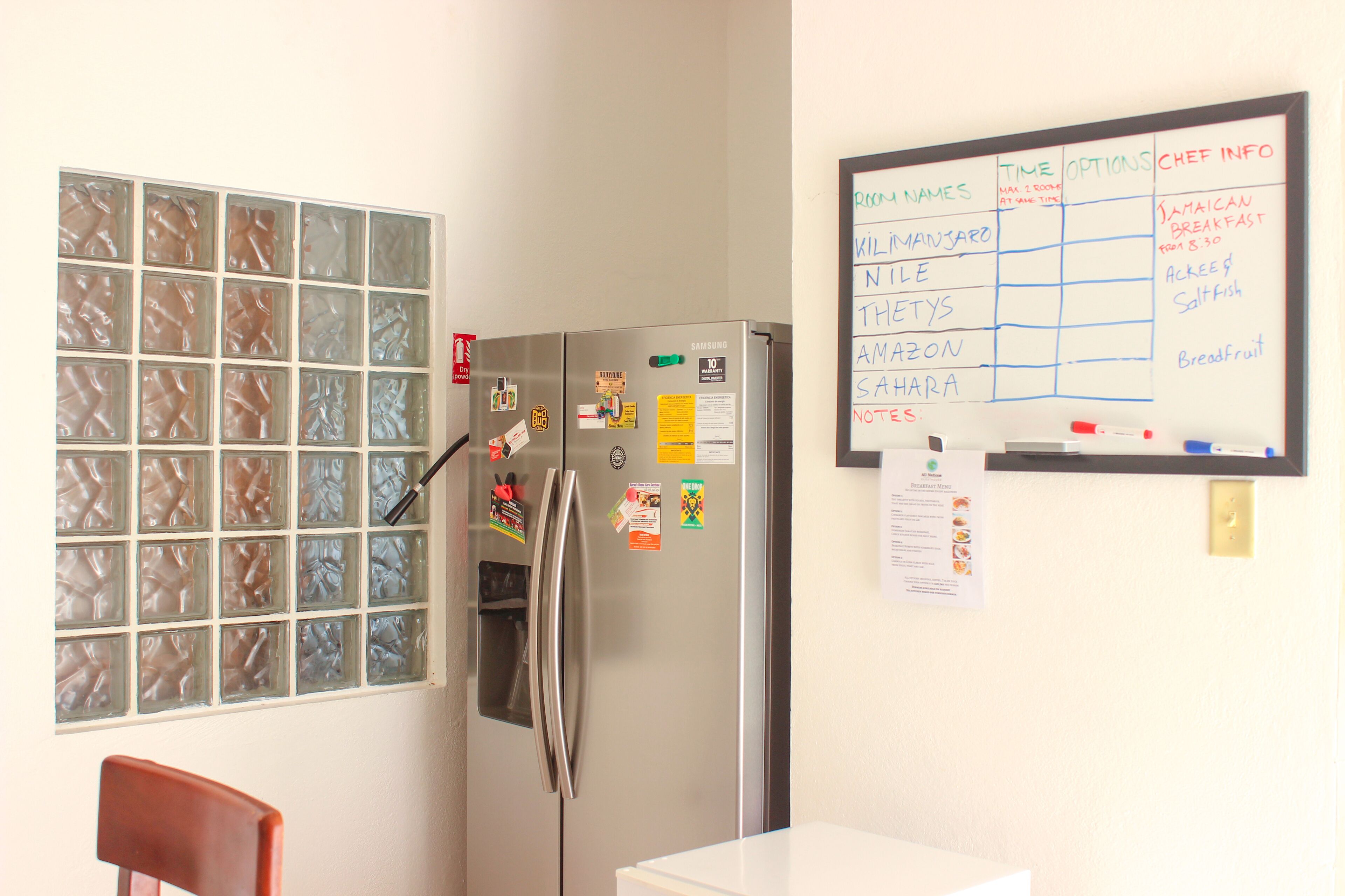 A kitchen with a stainless steel refrigerator, a whiteboard with room names and chef info, and a window with frosted glass panels.