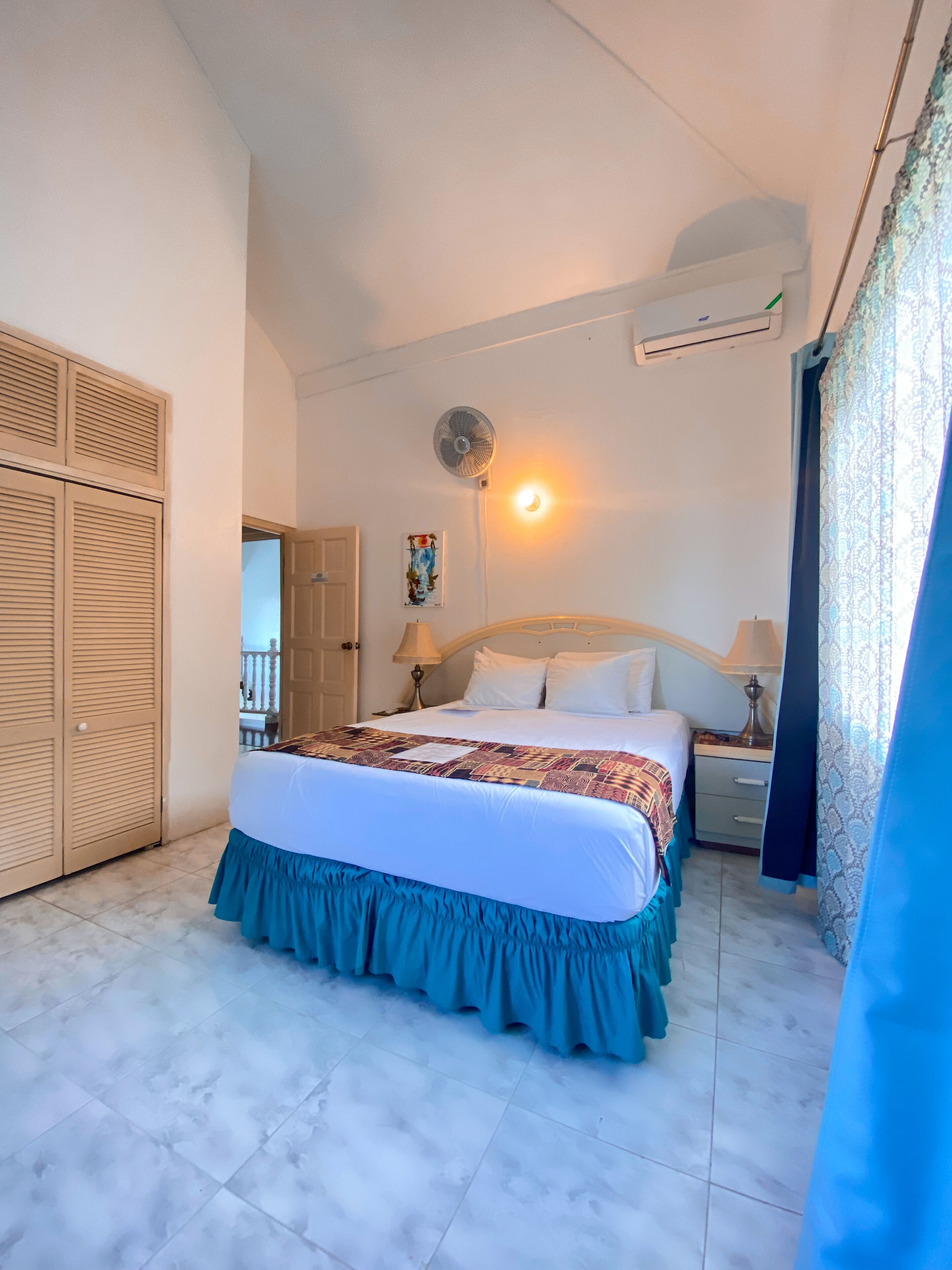 A bedroom with a bed, a bedside table, a lamp, a wall-mounted air conditioner, and a ceiling fan.