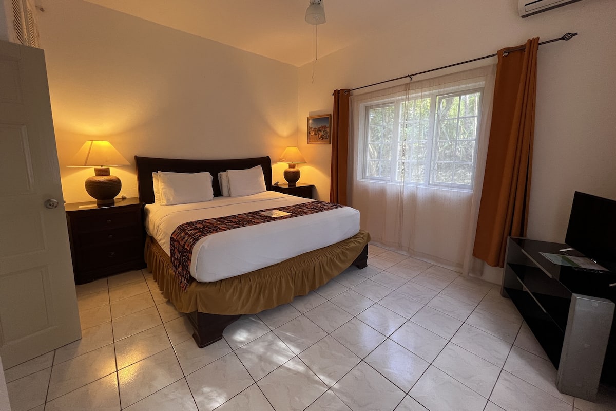 A hotel room with a bed, two bedside tables with lamps, a ceiling fan, a window with curtains, and a TV stand.
