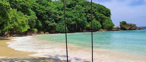 A beach with a swing overlooking turquoise waters and a forested hill.