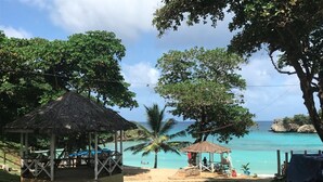 A beach area with two thatched-roof structures, one with a blue table and chairs, and the other with a single chair. - All Nations Guesthouse (Port Antonio)