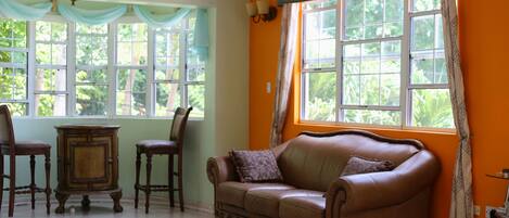 Una habitación con un sofá de cuero marrón, dos sillas de madera, una mesita de madera y un ventanal con cortinas.