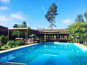 A swimming pool with a covered seating area and a building with a tiled roof in the background. - Hotel Hoja de Parra (Santa Cruz)