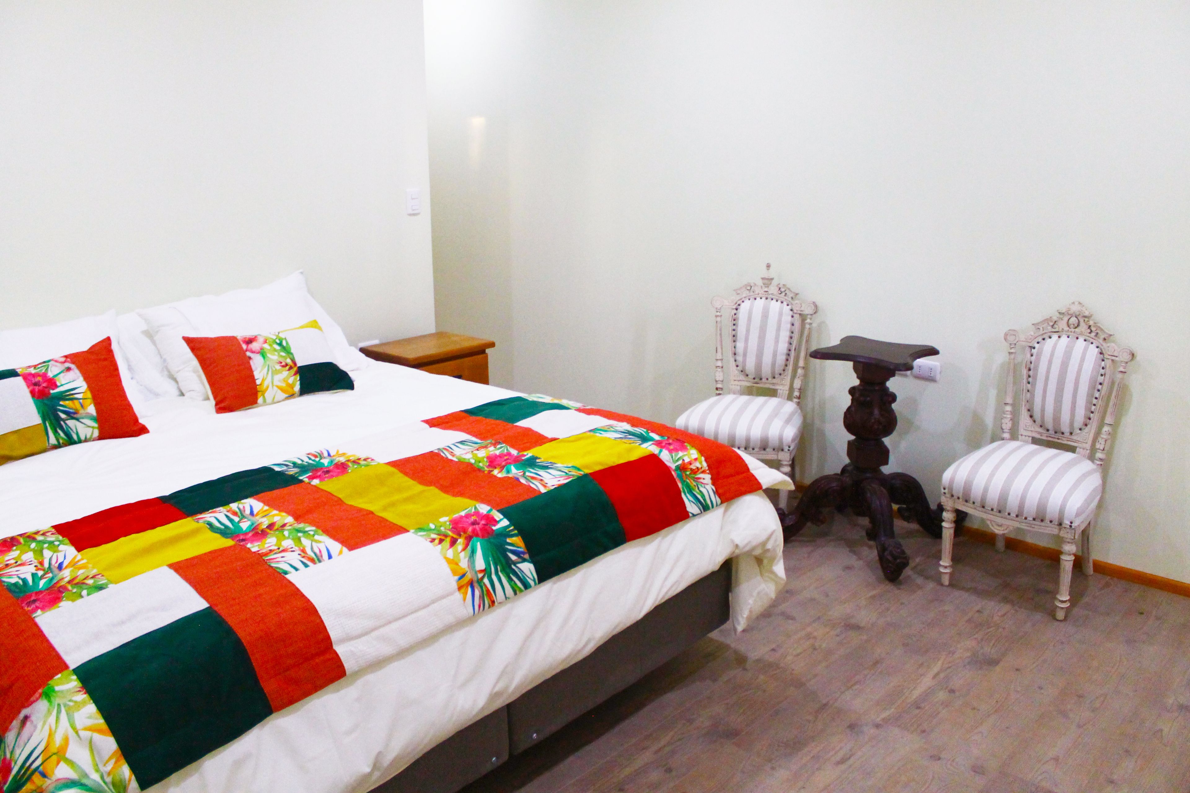 A neatly made bed with a colorful quilt, two upholstered chairs, a small table, and a wooden nightstand in a room with wooden flooring.