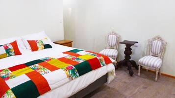 A neatly made bed with a colorful quilt, two upholstered chairs, a small table, and a wooden nightstand in a room with wooden flooring.