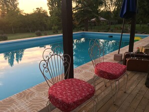 Zona junto a la piscina con dos sillas y una mesa, rodeada por una terraza de madera y una alberca.